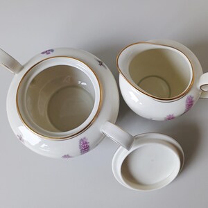 May include: A white porcelain tea set with gold trim, featuring a teapot, creamer, and lid. The set is decorated with purple floral patterns. The teapot has a handle and spout, and the creamer has a handle.