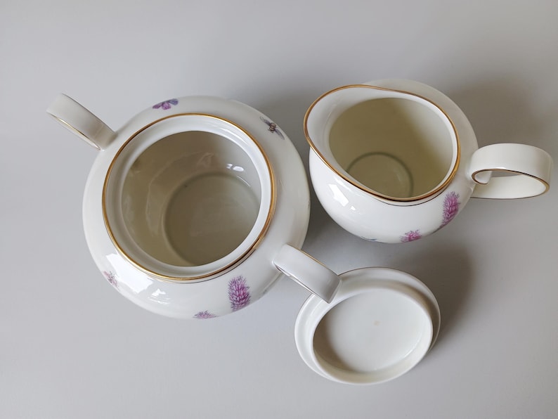 May include: A white porcelain tea set with gold trim, featuring a teapot, creamer, and lid. The set is decorated with purple floral patterns. The teapot has a handle and spout, and the creamer has a handle.