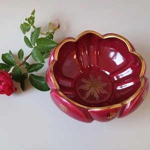May include: A deep red, scalloped glass bowl featuring a gold-trimmed rim and a gold starburst pattern at the center. The exterior of the bowl is decorated with small gold fan-shaped motifs. A single red rose with green leaves lies beside the bowl.