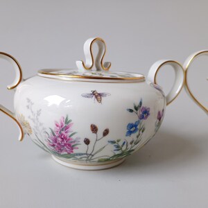 May include: A white porcelain sugar bowl with a lid and matching creamer. The set is decorated with colorful floral and bee motifs, and gold trim. The sugar bowl has a handle on each side and a lid with a loop handle.
