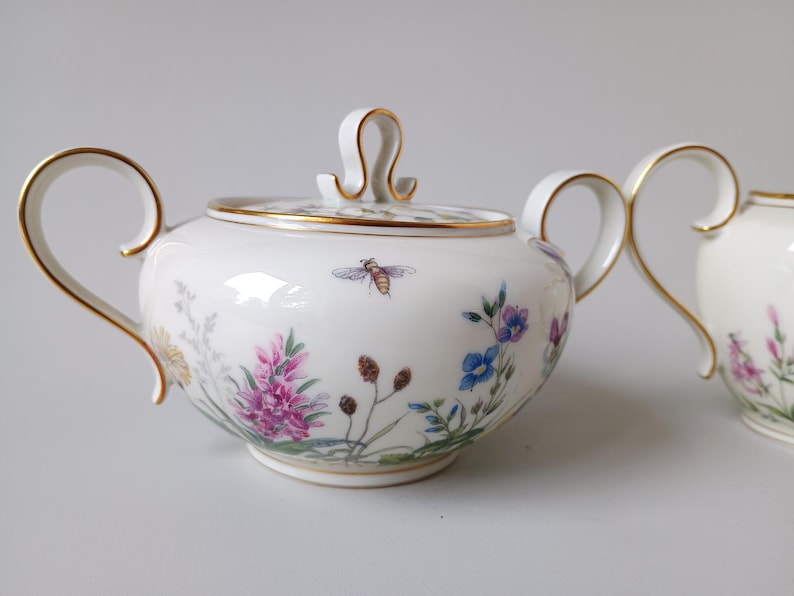 May include: A white porcelain sugar bowl with a lid and matching creamer. The set is decorated with colorful floral and bee motifs, and gold trim. The sugar bowl has a handle on each side and a lid with a loop handle.