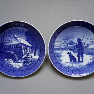 Puede incluir: Dos platos decorativos azules y blancos. Uno representa un molino de agua en un paisaje nevado, el otro un cazador con un perro. Ambos platos tienen un patr&oacute;n de estrellas y los a&ntilde;os 1976 y 1977.