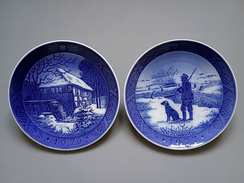 Puede incluir: Dos platos decorativos azules y blancos. Uno representa un molino de agua en un paisaje nevado, el otro un cazador con un perro. Ambos platos tienen un patr&oacute;n de estrellas y los a&ntilde;os 1976 y 1977.
