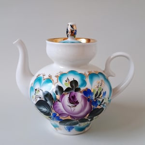 May include: A white porcelain teapot featuring a hand-painted floral design with a large central purple rose, dark green leaves, and bright turquoise accents. The teapot includes a matching lid with a gold-trimmed rim and a gold-painted handle.