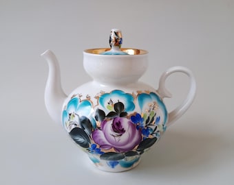Può includere: Una teiera in porcellana bianca con un motivo floreale dipinto a mano, caratterizzata da una grande rosa viola centrale, foglie verde scuro e vivaci accenti turchesi. Include un coperchio abbinato con bordo dorato e un manico dorato.