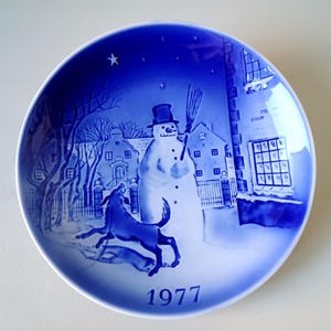 May include: A blue and white ceramic plate with a winter scene featuring a snowman, a dog, and houses. The plate has the year 1977 printed on the bottom.