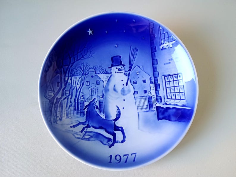 May include: A blue and white ceramic plate with a winter scene featuring a snowman, a dog, and houses. The plate has the year 1977 printed on the bottom.
