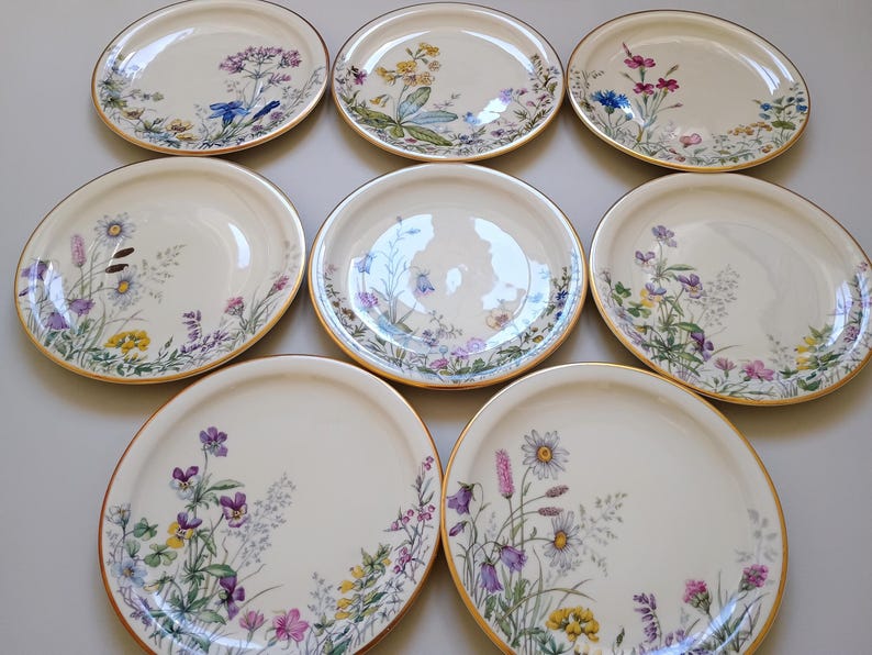 May include: Nine off-white ceramic plates with a gold rim, each decorated with a different floral design in shades of purple, yellow, and blue. The plates are arranged in a grid pattern, showcasing the variety of botanical illustrations.