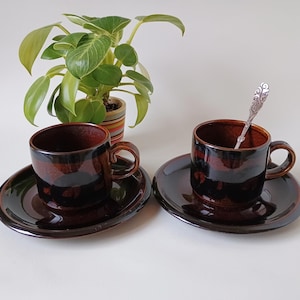 Könnte beinhalten: Zwei braune Espresso-Tassen und Untertassen aus Keramik mit glänzendem Finish. Eine Tasse hat einen dekorativen Löffel. Eine kleine Topfpflanze mit grünen Blättern befindet sich im Hintergrund.