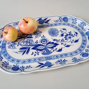 May include: An oval white ceramic serving platter with a scalloped edge, decorated with a blue floral and botanical pattern. Two round, pale yellow and pink apples rest on the left side of the platter, contrasting with the blue design.