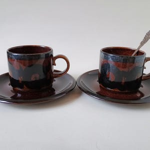 May include: Two ceramic espresso cups with matching saucers. The cups and saucers are dark brown with a glossy finish and a mottled pattern of dark brown and black. One cup has a small silver spoon.