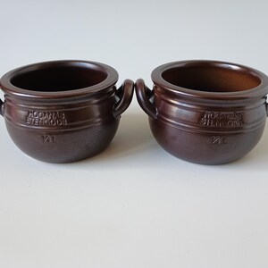 Può includere: Due pentole in ceramica marrone scuro con due manici laterali ciascuna. Le pentole presentano il testo in rilievo HÖGANÄS STENGODS e 1/2 L sulla parte anteriore. Questi recipienti rotondi a bocca larga hanno una finitura smaltata e liscia.
