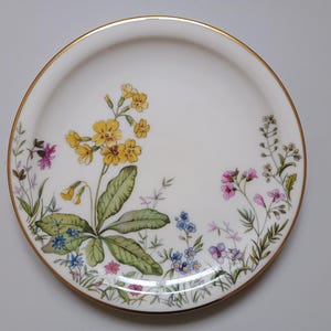 May include: White ceramic plate with a gold rim, decorated with a floral design. The flowers are yellow, pink, blue, and purple, with green leaves. A decorative piece for serving or display.