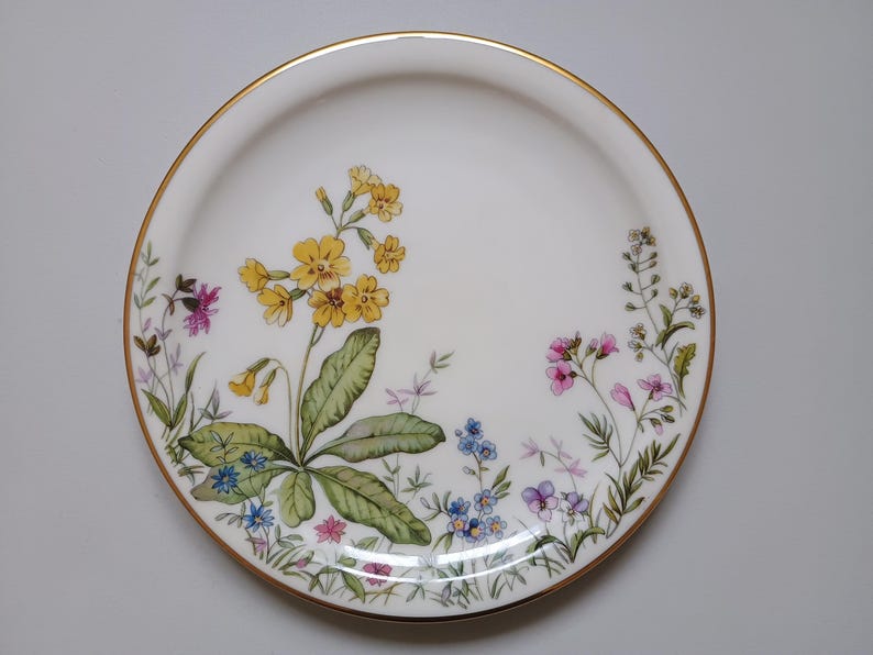 May include: White ceramic plate with a gold rim, decorated with a floral design. The flowers are yellow, pink, blue, and purple, with green leaves. A decorative piece for serving or display.