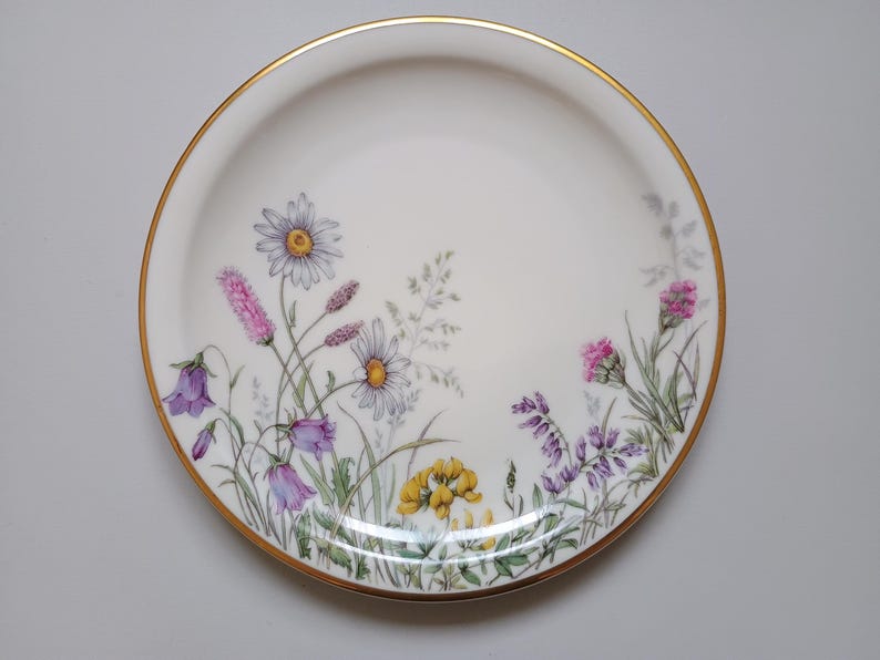 May include: A white ceramic plate with a gold rim, decorated with a floral design. The flowers include daisies, purple bellflowers, and pink and yellow wildflowers. The plate is a decorative piece for serving food.