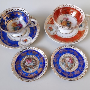 Op de afbeelding: Twee porseleinen theekopjes met bijpassende schoteltjes, één in kobaltblauw en één in oranje, beide met goudfiligraan, bloemmotieven en geschulpte randen. Twee extra kleine blauw-gouden decoratieve bordjes met centrale figuratieve scènes.