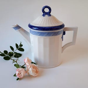 May include: A white porcelain teapot with vertical fluting, featuring a blue patterned border and a matching lid with a ring-shaped handle. A small sprig of three pale pink roses with green leaves is positioned next to the teapot base.