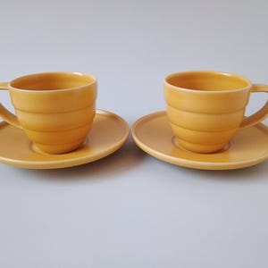 Puede incluir: Dos tazas de té y platillos de cerámica amarillos a juego. Las tazas tienen un diseño acanalado y asas curvas. Los platillos son redondos y planos. El juego es de un color cálido y acogedor.