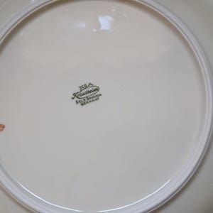 May include: Cream-colored ceramic plate with a white rim. The plate has a glossy finish and a decorative border. The bottom of the plate features a green stamp that reads "K.B.A. Krautheim Selb Bavaria Germany."