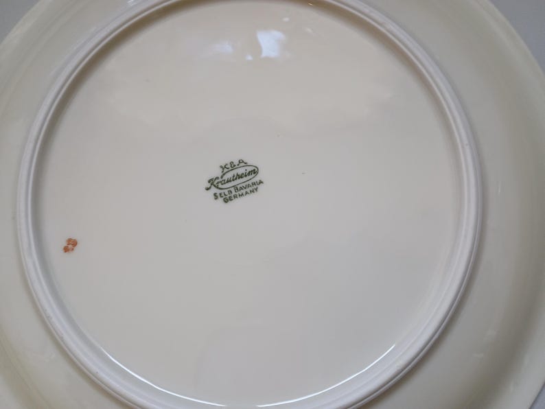 May include: Cream-colored ceramic plate with a white rim. The plate has a glossy finish and a decorative border. The bottom of the plate features a green stamp that reads "K.B.A. Krautheim Selb Bavaria Germany."