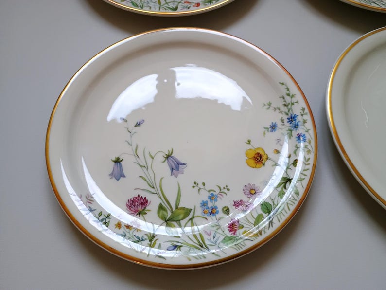 May include: A set of white ceramic plates with a gold rim and a floral design. The plates feature a variety of colorful flowers, including bluebells, yellow buttercups, and pink clover. The plates are likely for serving food.