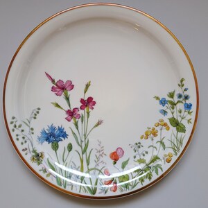 May include: White ceramic plate with a gold rim, decorated with colorful wildflowers. The floral design includes pink, blue, yellow, and orange blossoms with green foliage. A decorative piece for serving or display.