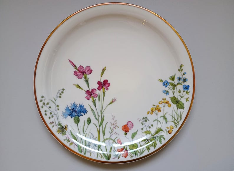 May include: White ceramic plate with a gold rim, decorated with colorful wildflowers. The floral design includes pink, blue, yellow, and orange blossoms with green foliage. A decorative piece for serving or display.