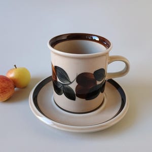 Puede incluir: Un juego de taza y platillo de cerámica en tono beige claro con borde marrón oscuro. La taza presenta un motivo botánico pintado de hojas y frutos en marrón oscuro y negro. Dos manzanas pequeñas están colocadas a la izquierda del platillo.
