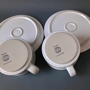 K&ouml;nnte beinhalten: Zwei wei&szlig;e Keramik-Teetassen und Untertassen mit einem schwarzen Logo, das auf der Unterseite der Tassen "Stoneware" und "Simply Home" lautet.