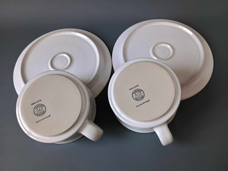 K&ouml;nnte beinhalten: Zwei wei&szlig;e Keramik-Teetassen und Untertassen mit einem schwarzen Logo, das auf der Unterseite der Tassen "Stoneware" und "Simply Home" lautet.