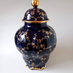 May include: A deep navy blue ceramic ginger jar with a matching lid, featuring a delicate gold floral vine pattern. The jar has a rounded body, a scalloped base, and a gold-finished finial on top, with gold trim along the lid and base edges.