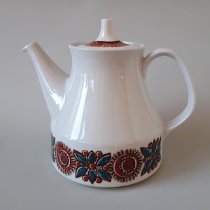 Op de afbeelding: Een witte keramische theepot met een taps toelopend lichaam en een bijpassend deksel met een puntige knop. De basis en het deksel zijn versierd met een herhalend bloemmotief in donkerrood, blauw en zwart, aangevuld met een gebogen handvat en tuit.