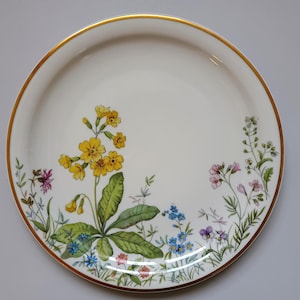 May include: A white ceramic plate with a gold rim, decorated with a floral design. The flowers include yellow primroses, blue forget-me-nots, and other colorful wildflowers. The plate is round.