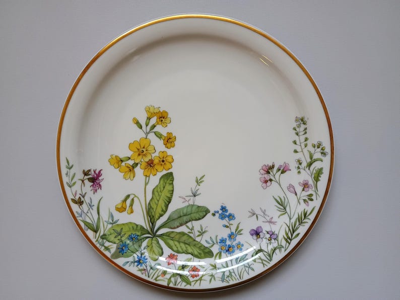 May include: A white ceramic plate with a gold rim, decorated with a floral design. The flowers include yellow primroses, blue forget-me-nots, and other colorful wildflowers. The plate is round.