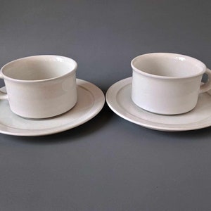 K&ouml;nnte beinhalten: Zwei wei&szlig;e Teetassen und Untertassen aus Keramik. Die Tassen haben ein schlichtes Design und eine leicht abgerundete Form. Die Untertassen haben einen d&uuml;nnen grauen Streifen am Rand.