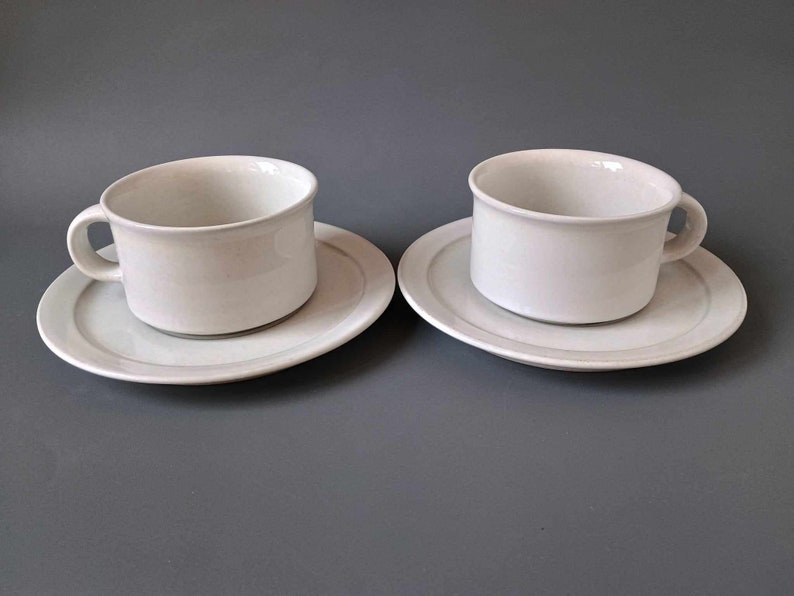 K&ouml;nnte beinhalten: Zwei wei&szlig;e Teetassen und Untertassen aus Keramik. Die Tassen haben ein schlichtes Design und eine leicht abgerundete Form. Die Untertassen haben einen d&uuml;nnen grauen Streifen am Rand.