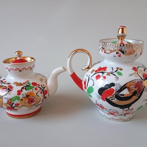 Pode incluir: Uma bule de porcelana branca e uma leiteira menor a combinar, ambos com tampas e pegas com acabamento dourado. Os recipientes estão decorados com ilustrações coloridas de galos, bagas vermelhas e folhas verdes, com detalhes em tinta dourada.