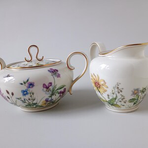 May include: A white porcelain sugar bowl with a lid and a creamer. Both pieces are decorated with floral patterns in purple, blue, yellow, and green, and have gold trim.