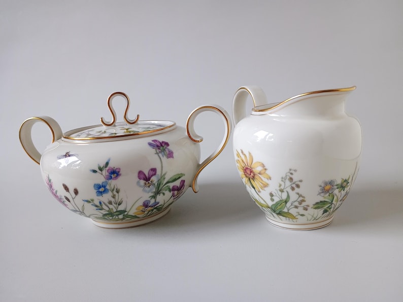 May include: A white porcelain sugar bowl with a lid and a creamer. Both pieces are decorated with floral patterns in purple, blue, yellow, and green, and have gold trim.