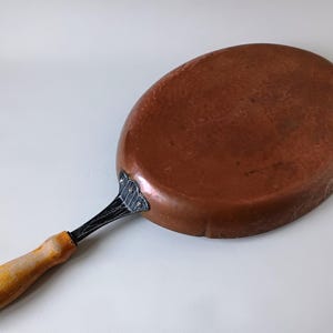 Könnte beinhalten: Eine ovale Kupferpfanne mit einer glatten, metallisch braunen Oberfläche. Die Pfanne verfügt über eine schwarze Metallgriffbefestigung mit drei sichtbaren Nieten, die mit einem hellen, zylindrischen Holzgriff mit abgerundetem Ende verbunden ist.