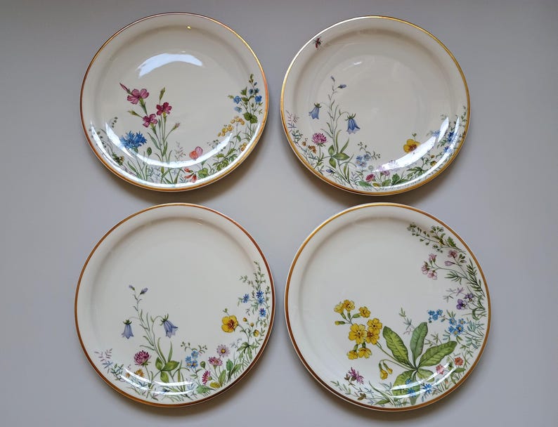 May include: Four white ceramic plates with a gold rim and floral designs. Each plate features a different arrangement of colorful wildflowers, including pink, blue, and yellow blossoms, creating a delicate and elegant aesthetic.
