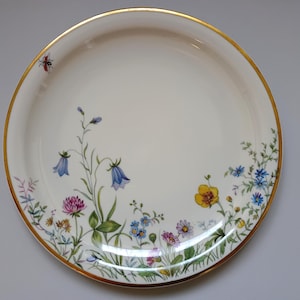 May include: A round, off-white ceramic plate with a gold rim. The plate is decorated with a floral design featuring bluebell, clover, and other wildflowers in shades of blue, yellow, pink, and green. A small red insect is near the rim.