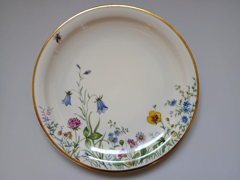 May include: A round, off-white ceramic plate with a gold rim. The plate is decorated with a floral design featuring bluebell, clover, and other wildflowers in shades of blue, yellow, pink, and green. A small red insect is near the rim.