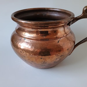 May include: A round, hammered copper pot with a wide, open top and a single curved handle attached to the side. The metal surface features a warm, metallic brown finish with visible texture from the hammering process and a smooth, rounded rim.