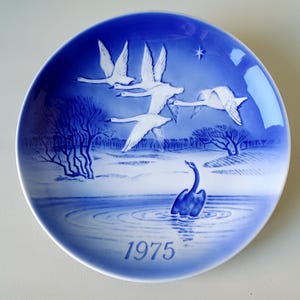 May include: A blue and white porcelain plate with a design of swans flying over a lake. The plate is part of a series and is dated 1975.