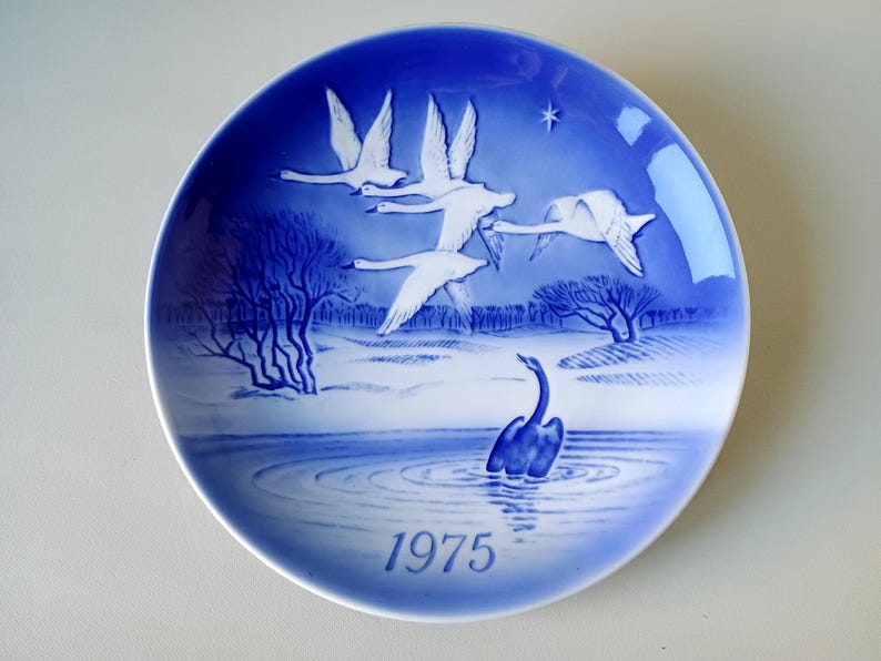 May include: A blue and white porcelain plate with a design of swans flying over a lake. The plate is part of a series and is dated 1975.