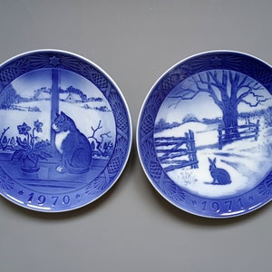 Puede incluir: Dos platos decorativos azules y blancos. Uno presenta un gato mirando por una ventana, el otro un conejo en una escena invernal. Ambos platos tienen un borde de estrella y los a&ntilde;os 1970 y 1971.