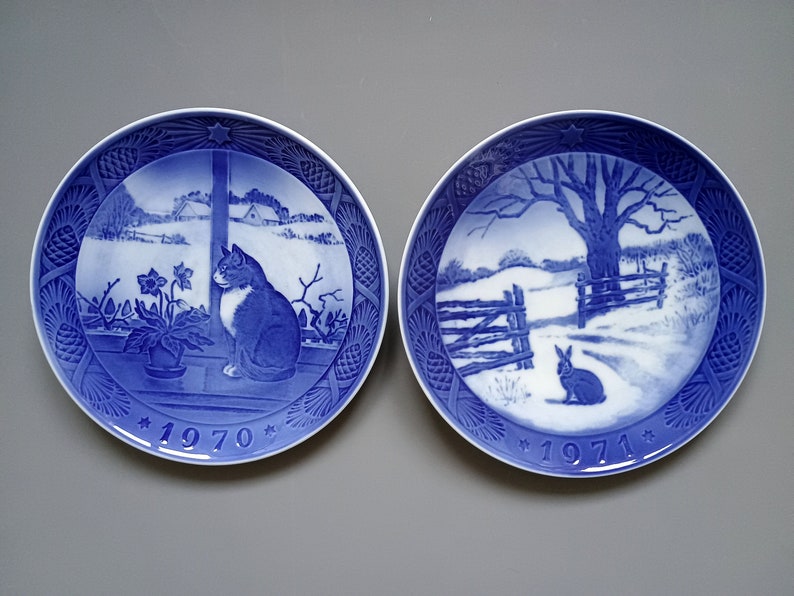 Puede incluir: Dos platos decorativos azules y blancos. Uno presenta un gato mirando por una ventana, el otro un conejo en una escena invernal. Ambos platos tienen un borde de estrella y los a&ntilde;os 1970 y 1971.