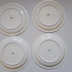 May include: Four white ceramic plates with a cream-colored rim and a decorative emblem on the underside. The plates are arranged in a square formation, showcasing their simple, elegant design. The plates are clean and appear to be in good condition.
