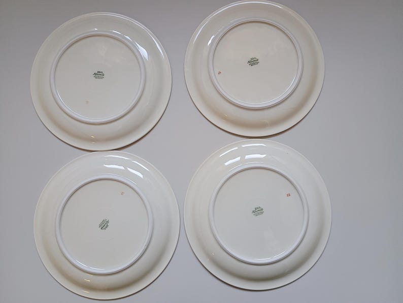 May include: Four white ceramic plates with a cream-colored rim and a decorative emblem on the underside. The plates are arranged in a square formation, showcasing their simple, elegant design. The plates are clean and appear to be in good condition.